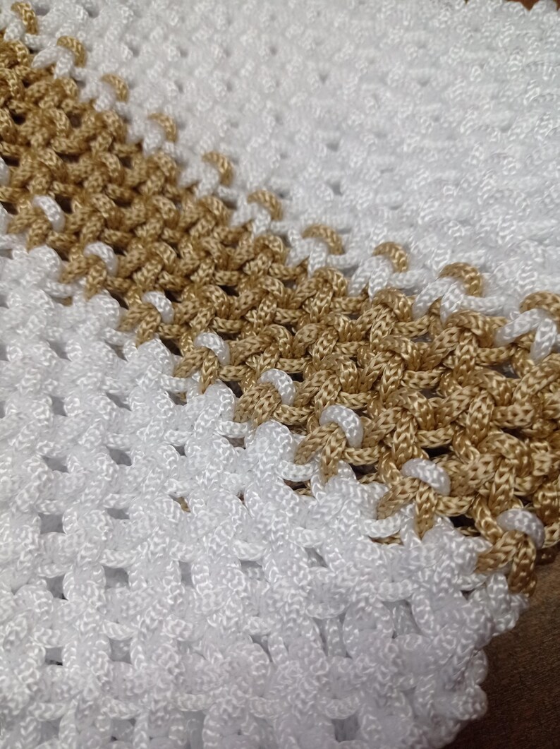 Puede incluir: Un art&iacute;culo textil con un cord&oacute;n blanco y dorado met&aacute;lico tejido en un patr&oacute;n de nudos repetitivos. El dise&ntilde;o consiste en una banda ancha de nudos blancos seguida de una franja diagonal de nudos dorados, creando una superficie de rejilla.