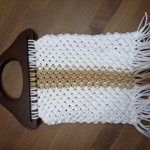 Puede incluir: Un bolso de macram&eacute; con un asa de pl&aacute;stico marr&oacute;n oscuro con una abertura circular. El cuerpo est&aacute; hecho de cord&oacute;n blanco con un patr&oacute;n de nudos en forma de diamante, acentuado por una franja central horizontal de cord&oacute;n dorado y flecos.