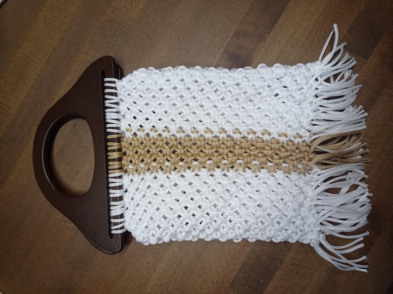 Puede incluir: Un bolso de macram&eacute; con un asa de pl&aacute;stico marr&oacute;n oscuro con una abertura circular. El cuerpo est&aacute; hecho de cord&oacute;n blanco con un patr&oacute;n de nudos en forma de diamante, acentuado por una franja central horizontal de cord&oacute;n dorado y flecos.