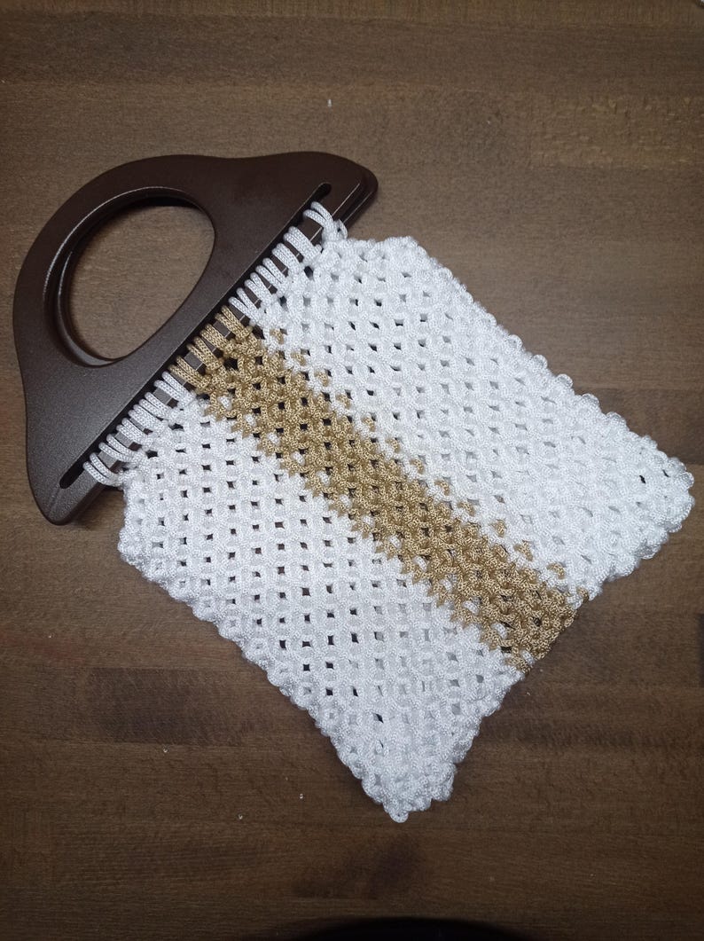 Puede incluir: Un bolso peque&ntilde;o con cuerpo de cord&oacute;n de macram&eacute; blanco y tostado con un patr&oacute;n de celos&iacute;a en forma de diamante. El bolso est&aacute; unido a un asa r&iacute;gida de pl&aacute;stico de color marr&oacute;n oscuro en forma de semic&iacute;rculo con un centro recortado.