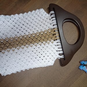 Puede incluir: Un bolso de macram&eacute; blanco y dorado con un tejido de patr&oacute;n de diamantes y un asa r&iacute;gida semicircular de color marr&oacute;n oscuro. Un peque&ntilde;o acento decorativo en forma de mariposa azul y negra se encuentra cerca de la parte inferior derecha.