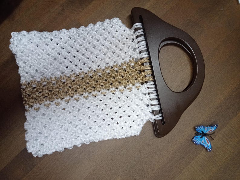 Puede incluir: Un bolso de macram&eacute; blanco y dorado con un tejido de patr&oacute;n de diamantes y un asa r&iacute;gida semicircular de color marr&oacute;n oscuro. Un peque&ntilde;o acento decorativo en forma de mariposa azul y negra se encuentra cerca de la parte inferior derecha.