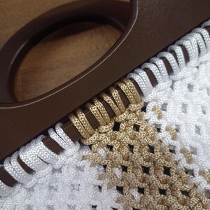 Puede incluir: Un bolso con un asa circular de color marr&oacute;n oscuro unida a un cuerpo de macram&eacute; blanco y beige. El cord&oacute;n est&aacute; enrollado alrededor del asa en un patr&oacute;n repetitivo, pasando de blanco a beige, creando un dise&ntilde;o de rejilla anudada y texturizada.