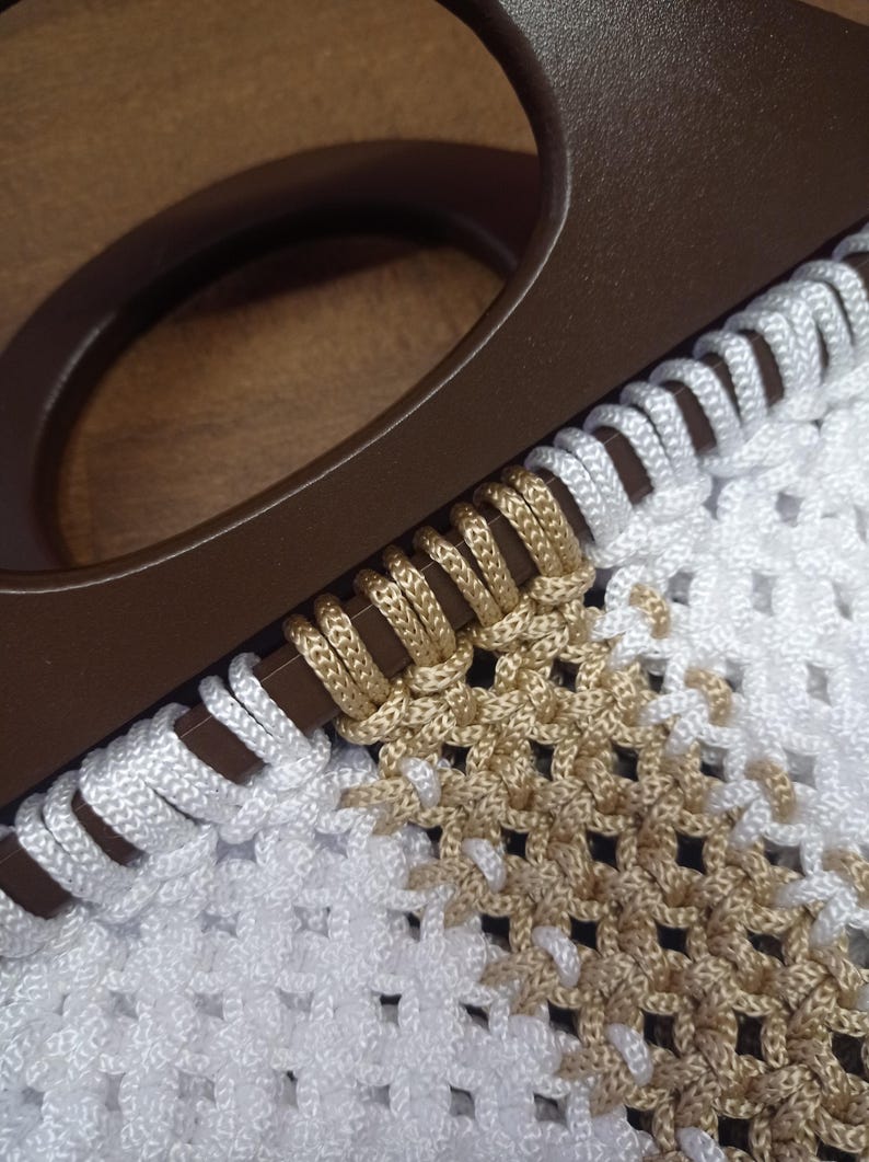 Puede incluir: Un bolso con un asa circular de color marr&oacute;n oscuro unida a un cuerpo de macram&eacute; blanco y beige. El cord&oacute;n est&aacute; enrollado alrededor del asa en un patr&oacute;n repetitivo, pasando de blanco a beige, creando un dise&ntilde;o de rejilla anudada y texturizada.