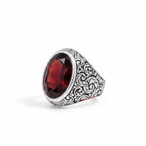Op de afbeelding: Een zilveren ring met een grote, ovaal geslepen dieprode edelsteen, gezet in een band met ingewikkelde, wervelende gegraveerde patronen. Het metalen oppervlak heeft een gepolijste afwerking met donkere uitsparingen in het ontwerp.