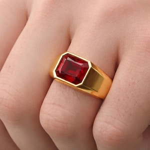May include: A gold ring featuring a large, rectangular, faceted red gemstone set in a smooth, polished gold band. The ring has a wide, solid metal shank that tapers slightly toward the back, showcasing a bold, clean design with a vibrant red stone.