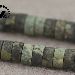 May include: Close-up of a strand of dark green and gray beads. The beads are cylindrical with a flat surface and a hole through the center. The beads have a mottled appearance, with variations in color and texture. The beads are strung together.