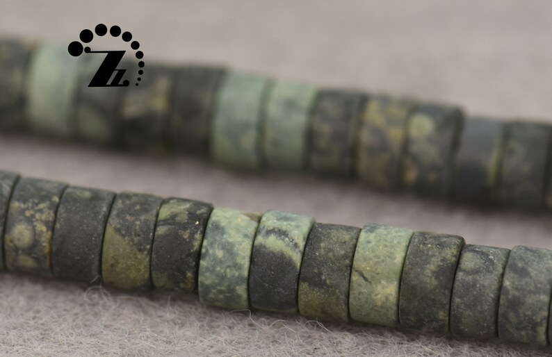 May include: Close-up of a strand of dark green and gray beads. The beads are cylindrical with a flat surface and a hole through the center. The beads have a mottled appearance, with variations in color and texture. The beads are strung together.