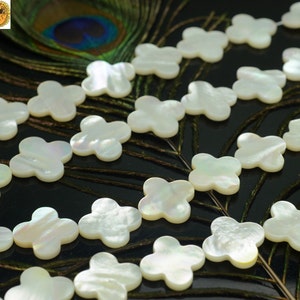 May include: A strand of white mother of pearl beads in the shape of four-leaf clovers. The beads are arranged in a line and are lying on a peacock feather.