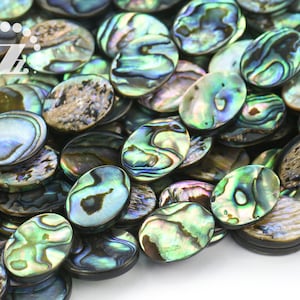 May include: A close-up shot of a pile of oval-shaped abalone shell beads. The beads display iridescent colors of green, blue, and purple, with unique swirling patterns. The shells have a smooth, polished surface.