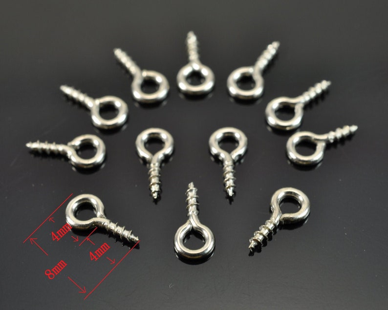 Puede incluir: Primer plano de 12 ganchos de ojo de tornillo de plata. Los ganchos tienen 8 mm de largo y un ojo de 4 mm de di&aacute;metro. Los ganchos est&aacute;n dispuestos en un c&iacute;rculo sobre una superficie negra.