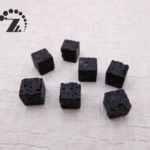 Puede incluir: Siete pequeños cubos de roca volcánica negra y porosa están dispersos sobre una superficie clara y texturizada. Los cubos tienen una textura rugosa y desigual y un color oscuro, carbón. La imagen presenta un logotipo en la esquina superior izquierda.