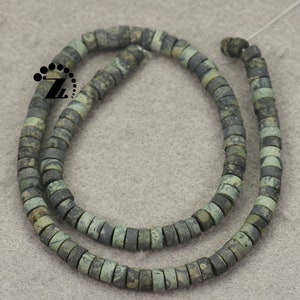 May include: Strands of flat, round beads in shades of green and gray. The beads have a mottled appearance, suggesting a natural stone material. The beads are strung together, forming a necklace or bracelet.
