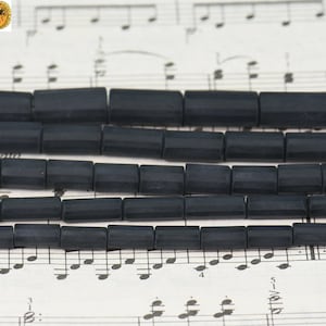 Può includere: Una collana di perline di vetro nero con forma cilindrica sfaccettata. Le perline sono disposte in file su un foglio di musica.