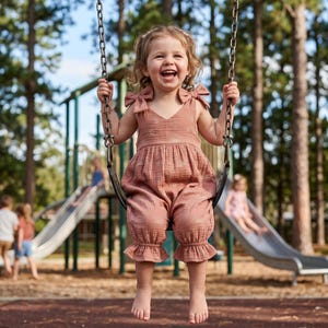 May include: A dusty rose-colored sleeveless romper featuring a V-neckline, shoulder ties, and ruffled leg openings. The garment is crafted from a textured fabric and has a loose, comfortable fit, suitable for active movement and outdoor play.