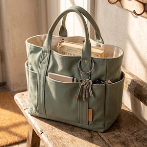 May include: A sage green canvas tote bag with two top handles, multiple exterior pockets, and a small rectangular brand tag. The bag contains a book titled Peter Wohlleben, a small notebook, and a metal keyring with several keys attached to it.