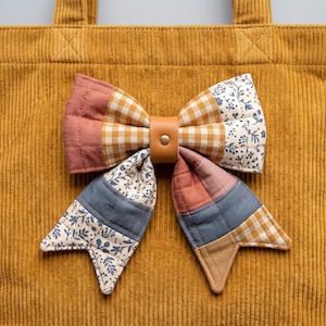 Op de afbeelding: Een mosterdgele corduroy tote bag met een grote, doorgestikte stoffen strik aan de voorzijde. De strik is opgebouwd uit patchwork-secties van effen oudroze, bruin-witte gingham en witte stof met blauwe bloemmotieven.