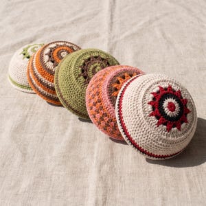 Puede incluir: Cinco kipás redondas de ganchillo dispuestas en fila. Cada una presenta un esquema de color único, incluyendo combinaciones de crema, naranja, marrón, verde oliva, rosa, rojo y negro, con intrincados patrones geométricos de estrellas.