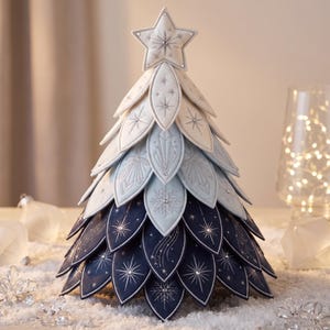 Op de afbeelding: Een decoratieve kerstboom voor op tafel, opgebouwd uit gelaagde, bloemblaadjesvormige stoffen elementen in een kleurverloop van wit naar lichtblauw en donker marineblauw. Elk blaadje heeft zilverkleurig borduurwerk van sterren en sneeuwvlokken.
