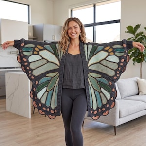 May include: A long-sleeved open-front cardigan featuring a large butterfly wing pattern in shades of dark gray, sage green, light blue, and soft orange. The garment has a ribbed trim along the front opening and cuffs, with a scalloped hemline.