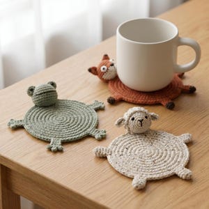 May include: Three round crochet coasters shaped like animals, including a sage green frog, a beige sheep, and a rust orange fox. The fox coaster supports a plain white ceramic mug. Each coaster features a circular base with four small limbs.