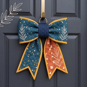 May include: A quilted fabric bow decoration featuring a teal and orange color palette with a geometric diamond pattern and white leaf line art. The bow has a central dark blue quilted knot and is suspended from a gold-colored metal door hook.