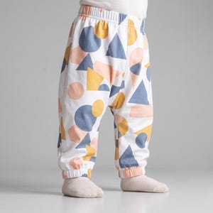 May include: White pants featuring a repeating geometric pattern of circles and triangles in shades of slate blue, mustard yellow, and soft peach. The garment has an elastic waistband and elasticized cuffs at the ankles, providing a gathered fit.