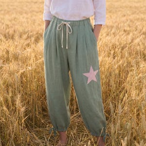 May include: Sage green trousers featuring a high-waisted design with a drawstring closure, pleated front detailing, and a single light pink star patch on the left leg. The trousers have gathered cuffs at the ankles and a relaxed, loose-fitting silhouette.