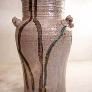 May include: A handmade ceramic vase with a textured, light brown surface. Dark brown and teal-colored lines cascade down the vase. The vase has two small handles and a wide, slightly flared rim. A unique piece for home decor.