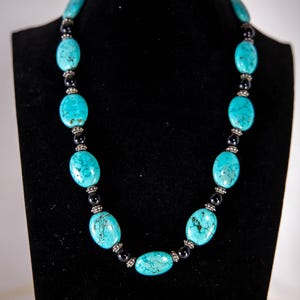May include: A necklace featuring oval-shaped turquoise stones with dark matrix patterns, interspersed with small, round, polished black beads and textured silver-toned metal spacers arranged in a repeating pattern along the length of the strand.