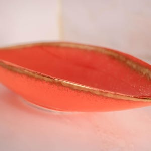 May include: An elongated, boat-shaped ceramic dish with a vibrant coral-orange interior and exterior. The rim features a textured, metallic gold-toned edge that contrasts with the smooth, matte finish of the bowl's body.