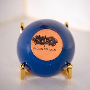 May include: A spherical blue ceramic object resting on a gold-toned metal stand. The base features a circular tan label with the text mud pie and MADE IN PORTUGAL printed in black. The object has a smooth, glossy finish and a rounded shape.