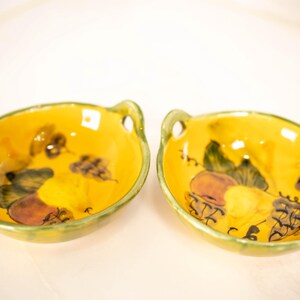 May include: Two oval-shaped yellow ceramic bowls with handles on each side. The interior of each bowl features a hand-painted design of green leaves and brown fruit. The edges of the bowls are finished with a green glaze, creating a distinct border.