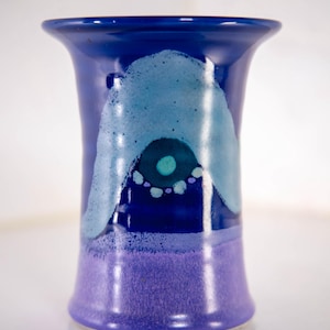 以下が含まれることがあります： 広がった縁を持つ陶器製の花瓶。上部は深い青色で、水色のアーチ状のデザインと、中央にティール色の点がある暗い円が描かれています。花瓶の下部は紫色の無地で、質感のある斑点状の仕上げが施されています。