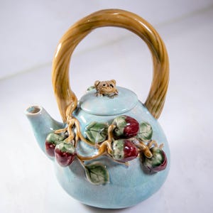 Può includere: Una teiera in ceramica azzurra con un grande manico ad arco e un piccolo beccuccio. La teiera è decorata con un ramo di frutta rossa e verde e foglie. Una piccola statuina di orso è seduta sul coperchio.