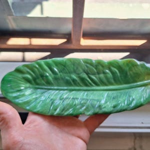 May include: A green, leaf-shaped decorative tray with a textured surface that mimics the veins and structure of a natural leaf. The object is elongated and pointed at one end, featuring a glossy finish with variations in green color tones.