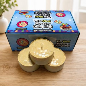 Op de afbeelding: Een blauwe kartonnen doos met het label The Viral Mystery Dumpling en illustraties van kleurrijke dumplings, geplaatst achter drie lichtbeige, ronde, geweven plastic bakjes met deksels, elk voorzien van een klein handvat in het midden.