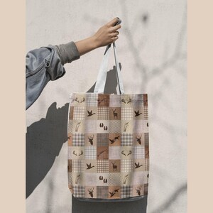 May include: A tote bag featuring a patchwork pattern of beige, brown, and white squares. The design includes repeating icons of deer antlers, silhouettes of deer, ducks in flight, hunting rifles, animal tracks, and tall grass stalks.