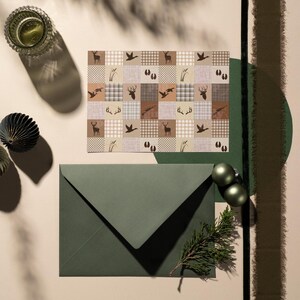May include: A rectangular card featuring a grid pattern of brown and beige squares with silhouettes of deer, ducks, antlers, tracks, and reeds, paired with a dark olive green envelope and a small sprig of evergreen needles.