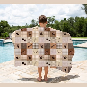 May include: A rectangular blanket featuring a patchwork pattern of brown and beige squares. The design includes repeating silhouettes of deer, antlers, ducks, animal tracks, and reeds, interspersed with solid colours and checkered patterns.
