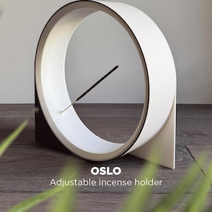 May include: A circular white incense holder with a ribbed exterior and a dark base. The object features the text OSLO Adjustable incense holder. A thin dark incense stick is positioned diagonally within the centre of the white circular frame.