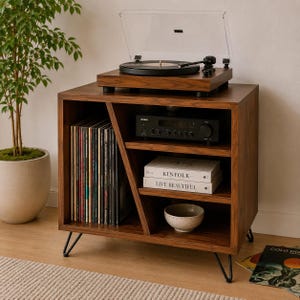 May include: A dark wood record player stand with black metal hairpin legs. The top holds a turntable. The interior features a vertical slot for vinyl records, a shelf with a black audio receiver, a shelf with two books titled Kinfolk and Live Beautiful, and a bottom shelf with a small ceramic bowl.