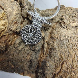May include: A silver-toned metal ring featuring a circular, hinged locket top. The face of the locket is decorated with an intricate, raised filigree pattern of swirling lines and small beads, centered around a single, small, polished silver sphere.