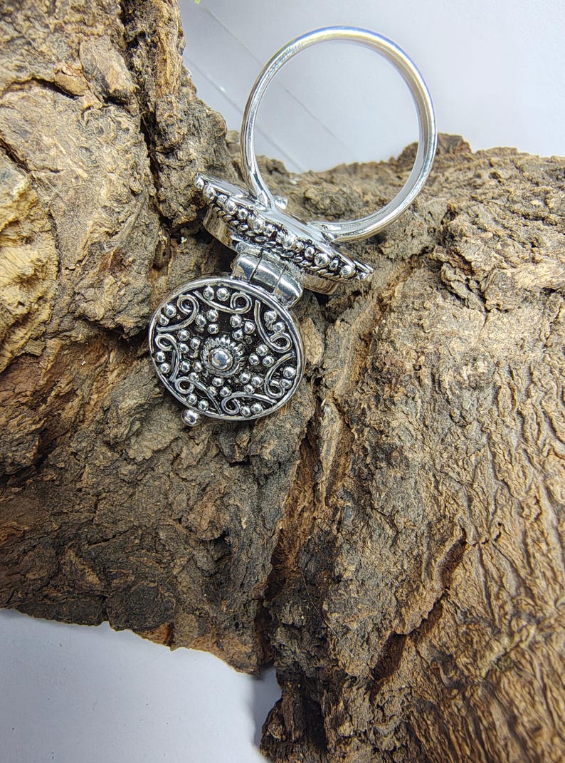 May include: A silver-toned metal ring featuring a circular, hinged locket top. The face of the locket is decorated with an intricate, raised filigree pattern of swirling lines and small beads, centered around a single, small, polished silver sphere.