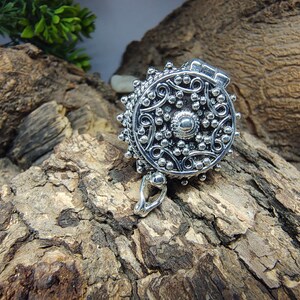 May include: A round silver locket pendant featuring an intricate embossed pattern of swirling lines and small raised dots. The circular metal piece has a hinged top section and a small silver bail at the bottom for attachment to a chain or cord.