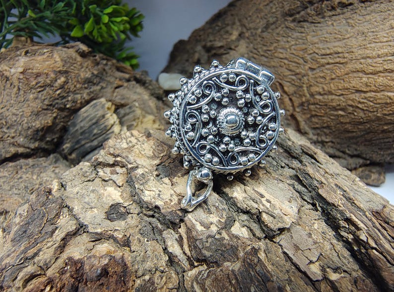 May include: A round silver locket pendant featuring an intricate embossed pattern of swirling lines and small raised dots. The circular metal piece has a hinged top section and a small silver bail at the bottom for attachment to a chain or cord.