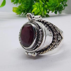 Può includere: Un anello medaglione in argento con una grande pietra preziosa sfaccettata di colore rosso scuro di forma ovale al centro. La fascia e l'incastonatura sono decorate con complesse texture a corda intrecciata, perline e una cerniera superiore.