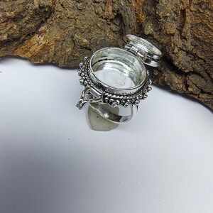 May include: A silver-toned metal ring featuring a circular, hinged locket compartment on the top. The locket is open, revealing a hollow interior. The outer edge of the circular setting is decorated with a series of small, raised metal beads.