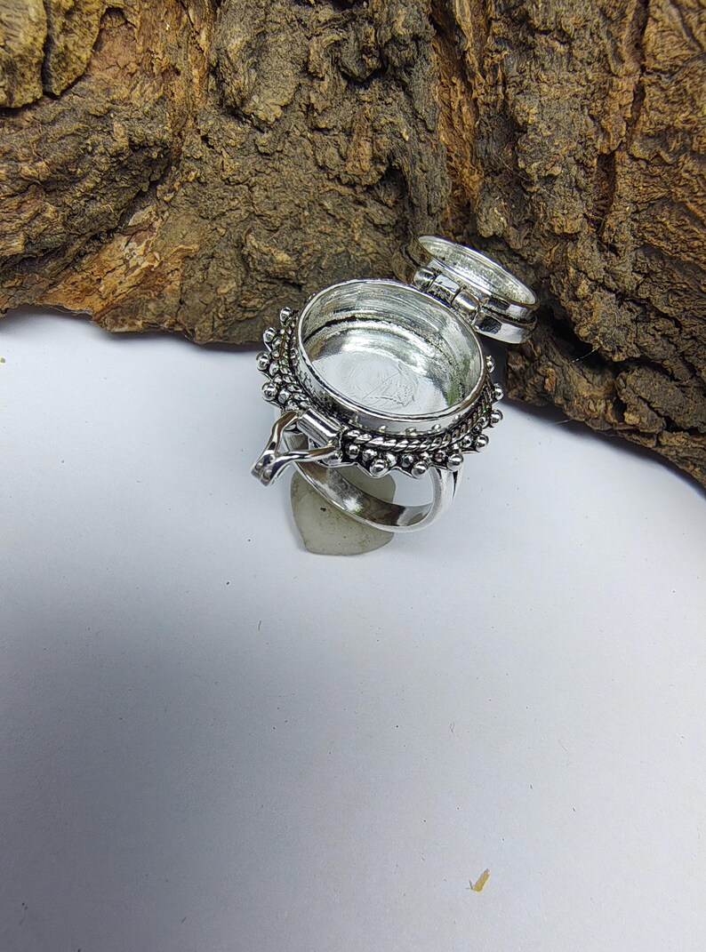 May include: A silver-toned metal ring featuring a circular, hinged locket compartment on the top. The locket is open, revealing a hollow interior. The outer edge of the circular setting is decorated with a series of small, raised metal beads.