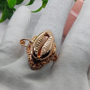 Può includere: Un anello color oro caratterizzato da un design centrale a forma di conchiglia di ciprea. La conchiglia è circondata da un bordo in metallo decorato in stile filigrana con intricati dettagli intagliati. L'intero pezzo ha una finitura lucida.